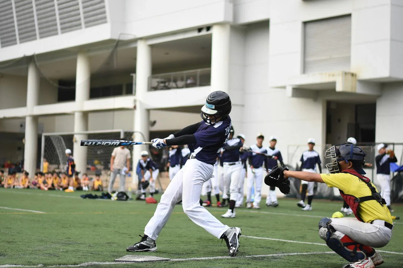 Youth baseball player swings a bat at a ball on a baseball field surrounded by teammates.