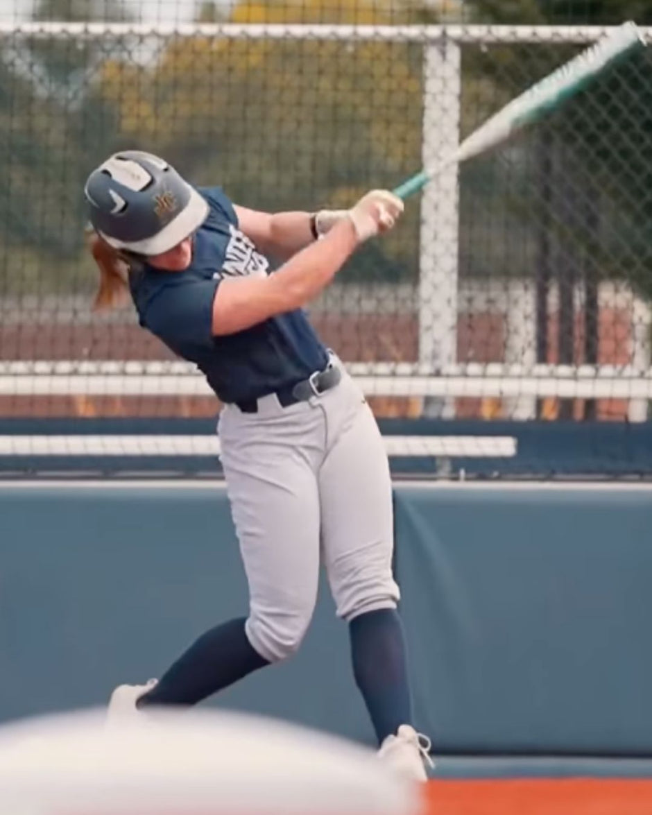 Person in a blue and gray softball uniform swinging a bat on a field.