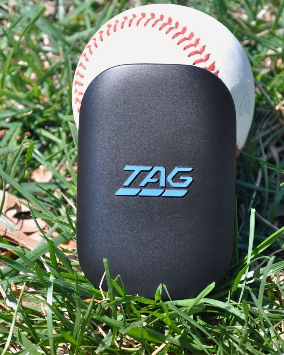 Black rectangular device with 'TAG' logo on grass next to a baseball