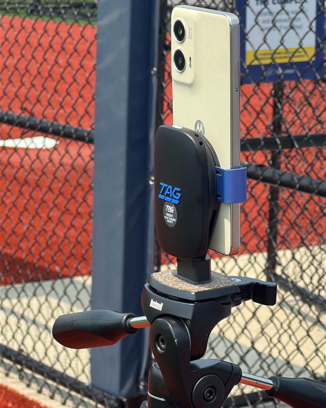 Phone holder with a phone attached, mounted on a sports equipment stand, with a chain-link fence and red surface in the background.