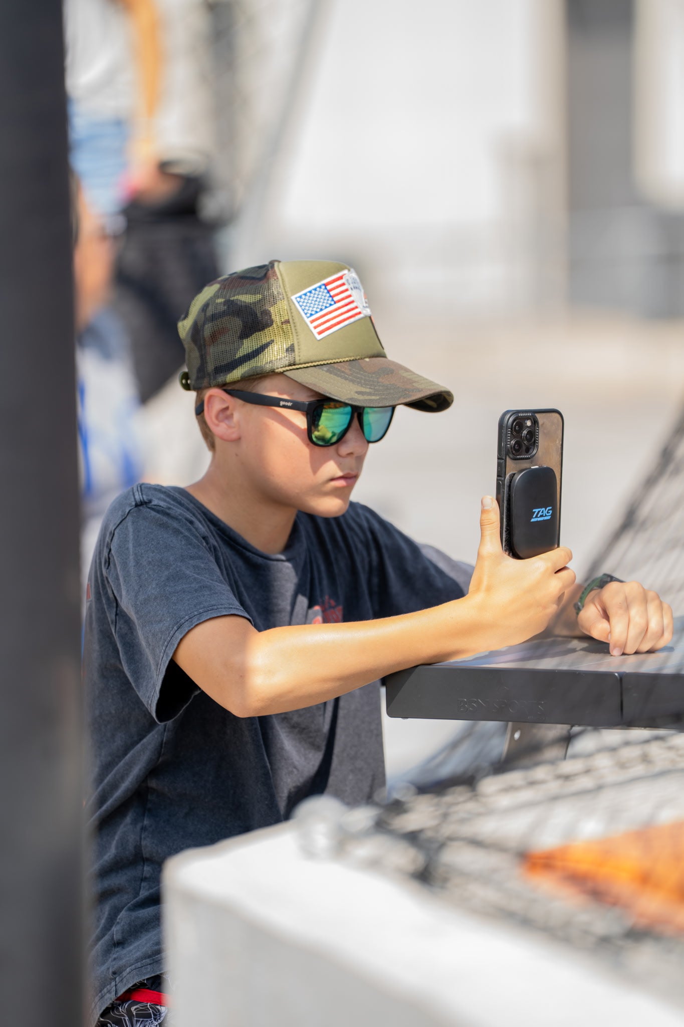 Person wearing a camouflage cap with an American flag patch, sunglasses, and a blue shirt, taking a photo with a smartphone.