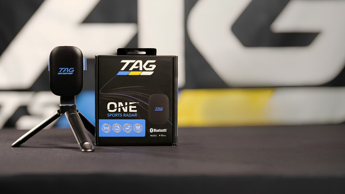 TAG ONE Sports Radar 2.4GHz wireless tracking device standing next to its retail packaging box on a dark surface, with a blurred TAG-branded background.