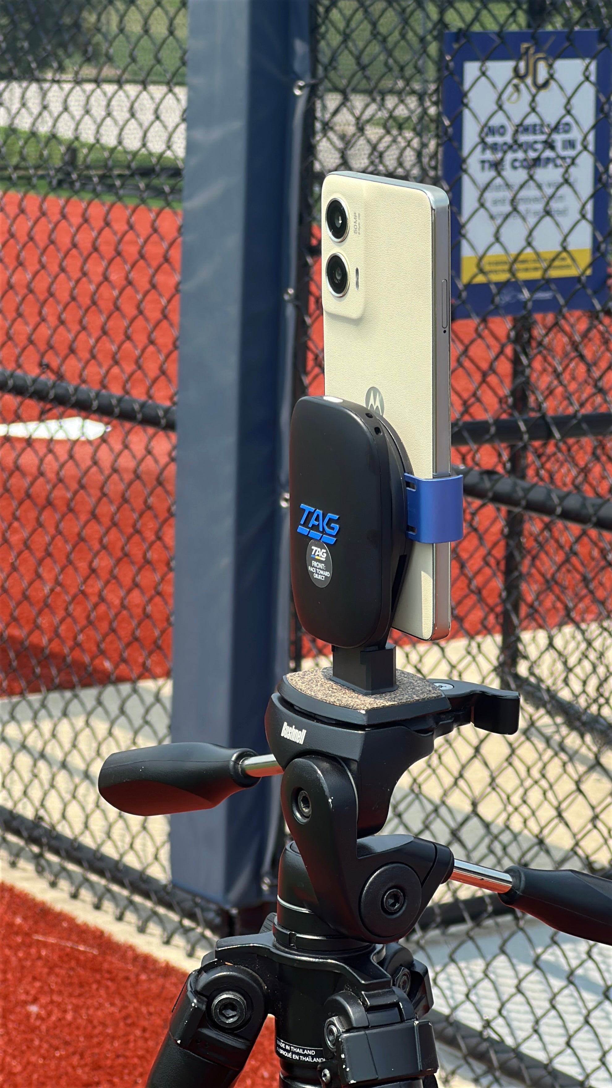 Phone mounted on a tripod with a TAG brand holder against a chain-link fence.