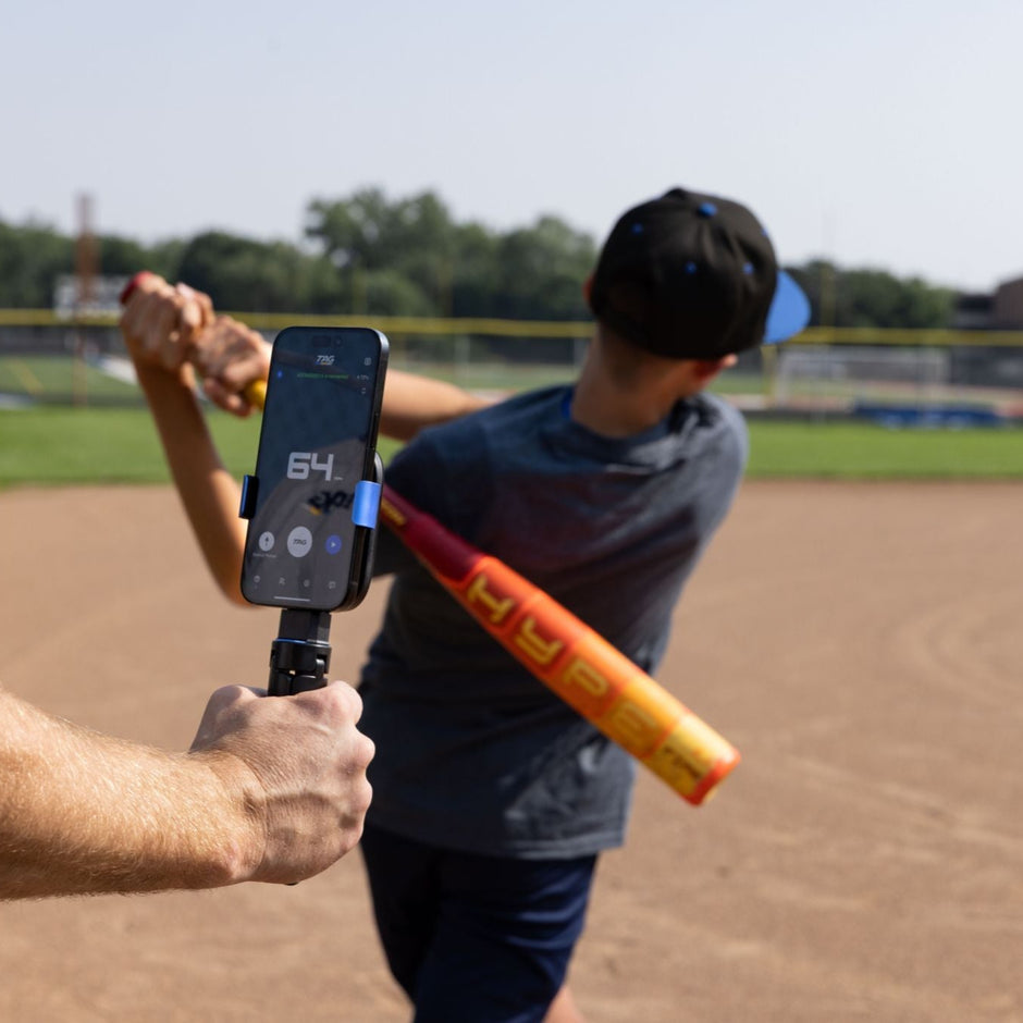 Youth baseball player swinging baseball bat while coach holds TAG One Sports Radar and tracks exit velocity of ball.