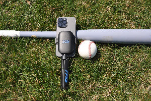 TAG One device with phone and tripod, resting on grass next to a baseball bat and ball.