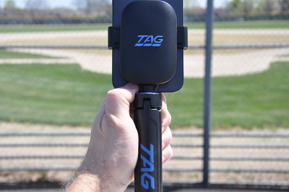 TAG One Sports Radar attached to bracket, phone, and tripod, held in user's hand in front of a baseball field.
